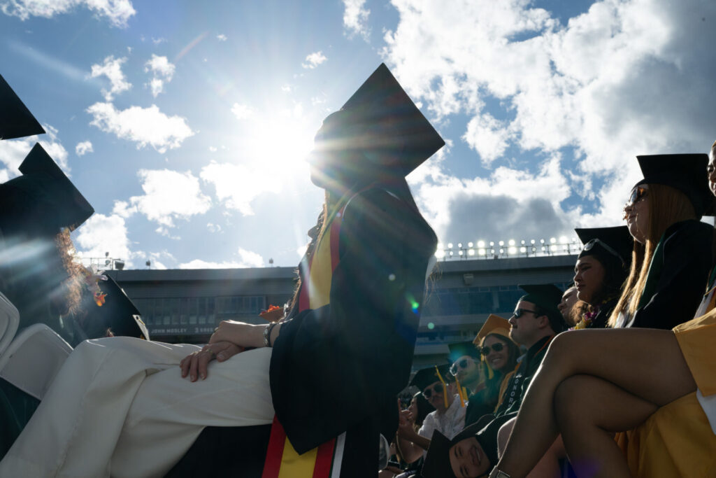 The camera looks up at a silhouette of a graduating student with the sun shining behind her.