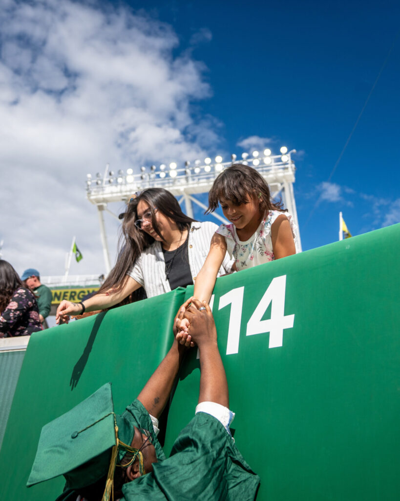 A graduating student reaches up to hold the hand of a child who is smiling down at him from the stands at Canvas Stadium.