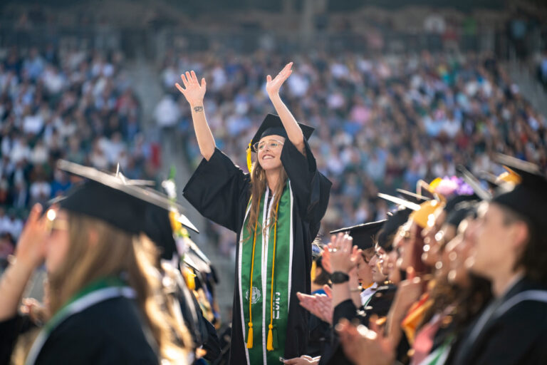 Graduating student stands among her peers on the Canvas Stadium field waving during Spring 2025 University-wide Commencement.