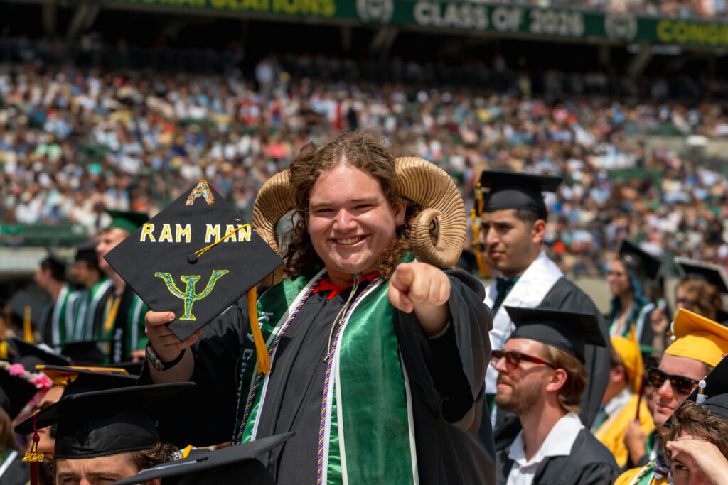 Graduating student wearing ram horns with his regalia - a signature look - smiles and points at the camera among the crowd at Canvas Stadium.